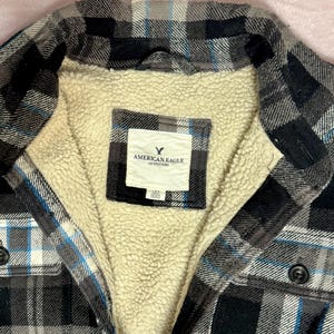 Puede incluir: Una camisa de franela a cuadros negros, grises y azules con forro de sherpa color crema. La camisa tiene una parte delantera abotonada y una etiqueta que dice "AMERICAN EAGLE OUTFITTERS."