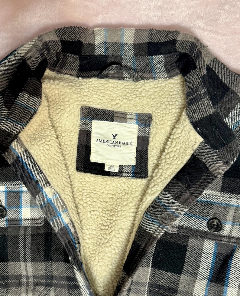 Puede incluir: Una camisa de franela a cuadros negros, grises y azules con forro de sherpa color crema. La camisa tiene una parte delantera abotonada y una etiqueta que dice "AMERICAN EAGLE OUTFITTERS."