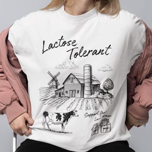 Pode incluir: Uma t-shirt branca de gola redonda com um gráfico a tinta preta de uma quinta com um celeiro, um moinho de vento e uma vaca. O texto "Lactose Tolerant" está no topo e "Support Your Local Farmer" na parte inferior, com dois jarros de leite.