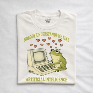 Pode incluir: Camiseta branca com um desenho de um sapo verde ao lado de um computador vintage. O texto diz "NINGUÉM ME ENTENDE COMO A INTELIGÊNCIA ARTIFICIAL" com ilustrações de corações.