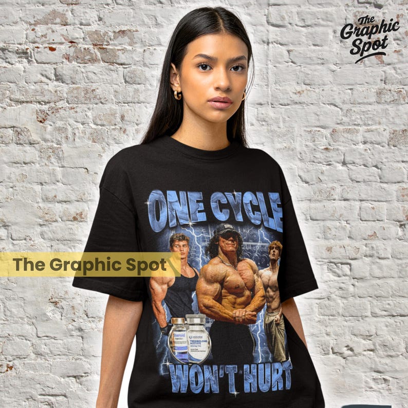 Woman wearing black oversized t-shirt with bold graphic reading One Cycle Wont Hurt, featuring illustrated bodybuilders, funny gym humor design