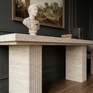 May include: A beige travertine console table with a rectangular top and two thick, vertically fluted pillar legs. A white stone bust of a Roman figure sits on the surface, accompanied by a stack of two dark-bound books on the right side.