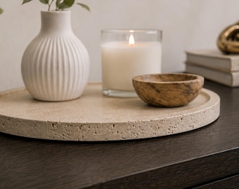 Op de afbeelding: Een rond, beige, poreus stenen dienblad bevat een witte geribbelde keramische vaas met groene bladeren, een brandende kaars in een heldere glazen pot en een kleine, ronde, bruine houten kom met een zichtbaar nerfpatroon, samen gerangschikt.