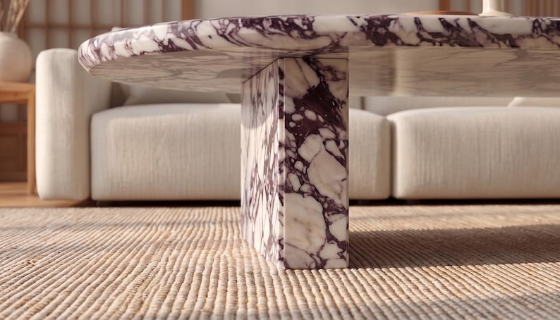 Op de afbeelding: Een ronde salontafel gemaakt van wit marmer met opvallende dieppaarse aders. De tafel heeft een dik, rond blad dat wordt ondersteund door een enkele, rechthoekige voet van hetzelfde bijpassende paars-witte marmermateriaal.