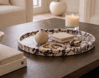 Op de afbeelding: Een rond dienblad met een wit en donkerpaars marmerpatroon. Op het dienblad staan een kleine beige vaas met droogbloemen, twee vierkante beige onderzetters, een snoer van houten kralen en een goudkleurige metalen knoopsculptuur.
