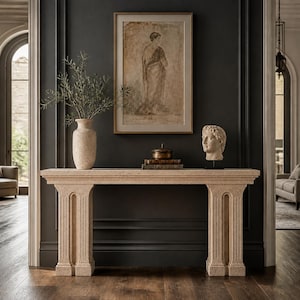 Peut inclure: Une console en pierre beige dotée de deux pieds en forme de colonnes cannelées. Sur la table se trouvent un grand vase moucheté avec des branches vertes, un petit récipient métallique à couvercle sur deux livres, et un buste en pierre blanche.