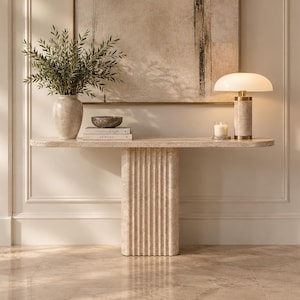 May include: A beige travertine console table with a smooth, elongated oval top and a central, vertically fluted pedestal base. The table features a porous, natural stone texture and stands against a wall, providing a surface for decorative items.