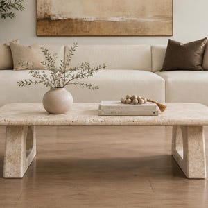 Peut inclure: Une table basse rectangulaire en pierre de travertin beige avec une surface poreuse et texturée et deux pieds en piédestal arqués. La table supporte un vase en céramique ronde avec des branches vertes, deux livres empilés et des perles en bois.