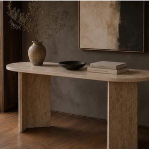 May include: An oval console table made of beige travertine stone with two thick, rectangular slab legs. On the table surface sits a textured brown ceramic vase with a branch, a shallow black bowl, and two stacked books with light-colored covers.