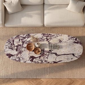 Op de afbeelding: Een ovale salontafel met een wit en donkerpaars marmeren blad met opvallende aders. De tafel staat centraal op een getextureerd beige tapijt en bevat twee kleine houten kommen en een klein rechthoekig glazen object op het oppervlak.