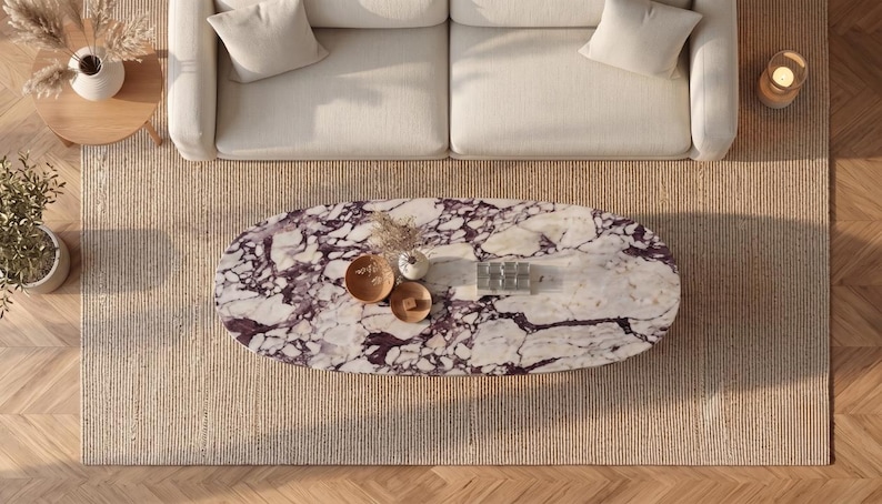 Op de afbeelding: Een ovale salontafel met een wit en donkerpaars marmeren blad met opvallende aders. De tafel staat centraal op een getextureerd beige tapijt en bevat twee kleine houten kommen en een klein rechthoekig glazen object op het oppervlak.