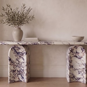 May include: A console table crafted from white marble with prominent purple veining, featuring a long, rounded rectangular top supported by two arched, thick marble legs. A ceramic vase with olive branches sits on the left, with books and a bowl.