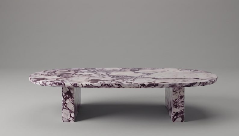 Op de afbeelding: Een ovale salontafel gemaakt van paars en wit geaderd marmer. De tafel heeft een glad, gepolijst bovenoppervlak met een opvallend brecciepatroon en wordt ondersteund door twee dikke, rechthoekige plaatpoten van hetzelfde steenmateriaal.
