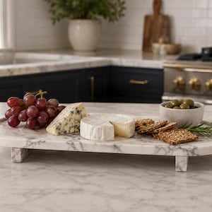 May include: An oval white marble serving tray with grey veining, supported by four short rectangular feet. The tray holds a cluster of red grapes, a wedge of blue cheese, a round wheel of brie, a small bowl of green olives, and several crackers.