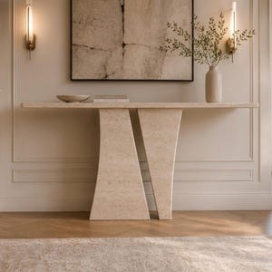 May include: A beige stone console table featuring a long, flat rectangular top supported by two distinct, angled pedestal bases. The table has a textured, porous surface and is positioned against a wall, holding a small bowl and a book.
