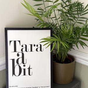 May include: A black framed print with the words "Tara a bit" in a bold, serif font. The text is accompanied by the definition: "(Ta-rah-a-bit) Friendly and informal way of saying "Goodbye" or "See you later". A potted plant is to the right.