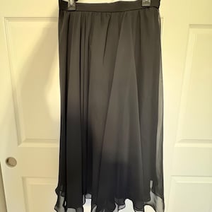 Puede incluir: Una falda larga y fluida de color negro cuelga de una percha negra. La falda est&aacute; hecha de una tela transparente y ligera que se drapea con gracia. El dobladillo tiene una sutil onda, que a&ntilde;ade movimiento al dise&ntilde;o. La falda se muestra contra una puerta blanca.