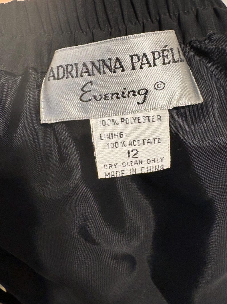 Puede incluir: Primer plano de una prenda negra con etiqueta. La etiqueta dice "ADRIANNA PAPELL Evening" y enumera los materiales como 100% poli&eacute;ster y forro 100% acetato. Tambi&eacute;n indica la talla 12 y "DRY CLEAN ONLY" con "MADE IN CHINA".