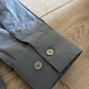 May include: Close-up of a gray dress shirt sleeve with three round buttons. The buttons are a light color with a decorative design. The fabric has a subtle texture.
