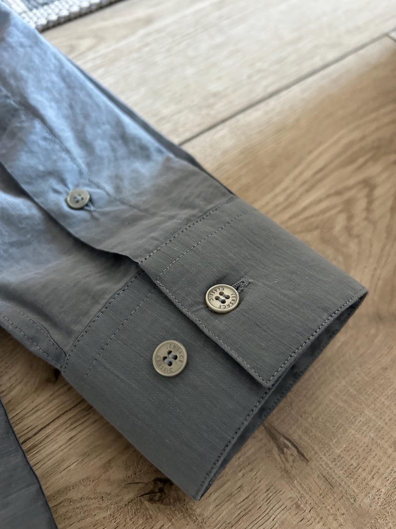 May include: Close-up of a gray dress shirt sleeve with three round buttons. The buttons are a light color with a decorative design. The fabric has a subtle texture.
