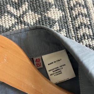May include: Close-up of a light blue shirt collar with a label. The label reads "MADE IN ITALY" and lists fabric composition: 60% cotton and 40% viscose. The shirt is hanging on a wooden hanger. The size is 40/151.