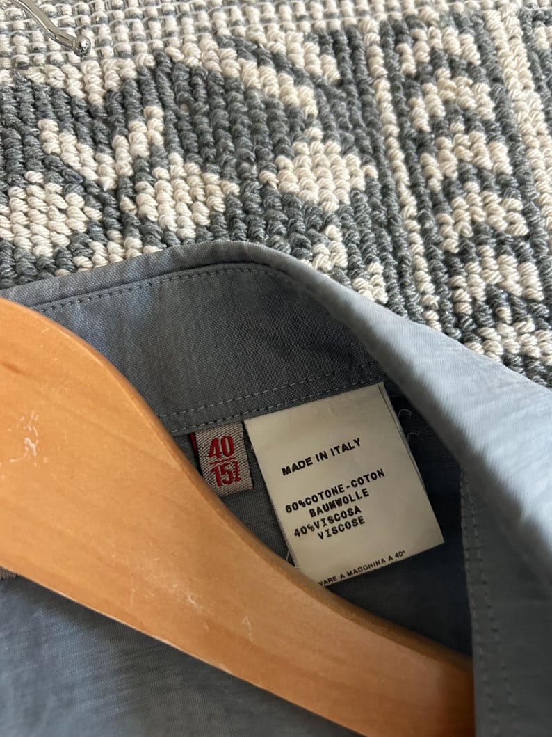 May include: Close-up of a light blue shirt collar with a label. The label reads "MADE IN ITALY" and lists fabric composition: 60% cotton and 40% viscose. The shirt is hanging on a wooden hanger. The size is 40/151.