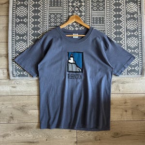 May include: A slate blue t-shirt featuring a graphic of a lighthouse with the word "WASHINGTON" printed below. The lighthouse graphic is framed in a blue square. The shirt is hanging on a wooden hanger.
