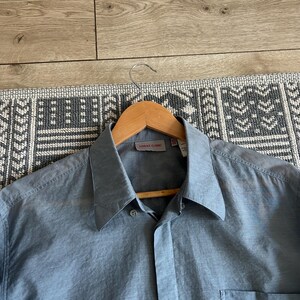 May include: A light blue button-down shirt with a classic collar and a chest pocket. The shirt is hanging on a wooden hanger. The label reads "VERSACE CLASSIC".
