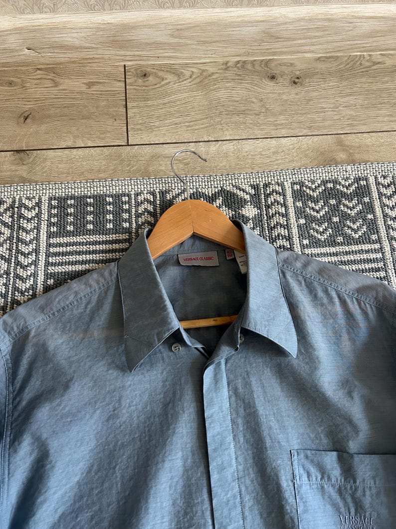 May include: A light blue button-down shirt with a classic collar and a chest pocket. The shirt is hanging on a wooden hanger. The label reads "VERSACE CLASSIC".