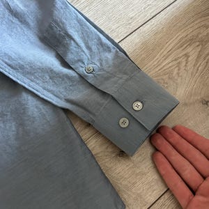 May include: Close-up of a light blue long-sleeve shirt. The shirt features a buttoned cuff with two buttons and a buttoned sleeve placket. The fabric has a subtle texture.