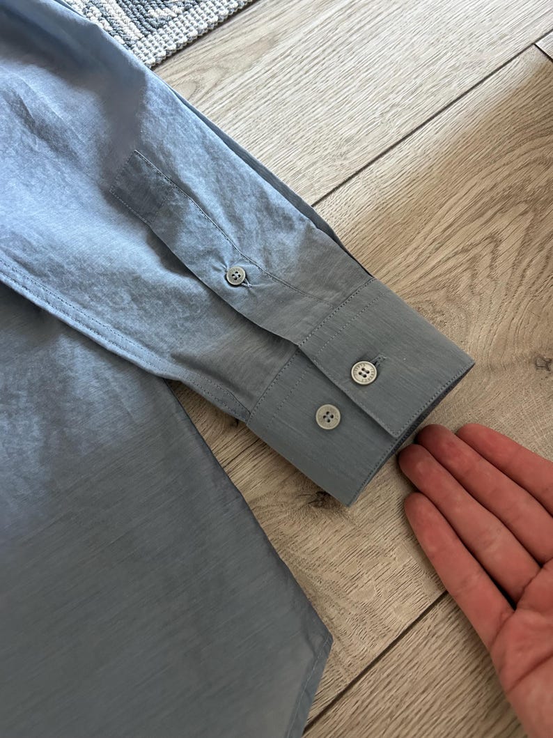 May include: Close-up of a light blue long-sleeve shirt. The shirt features a buttoned cuff with two buttons and a buttoned sleeve placket. The fabric has a subtle texture.