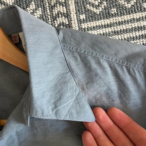 May include: Close-up of a light blue button-down shirt. The collar and part of the shirt are visible, with a tag showing size 40/15. The shirt is hanging on a wooden hanger.
