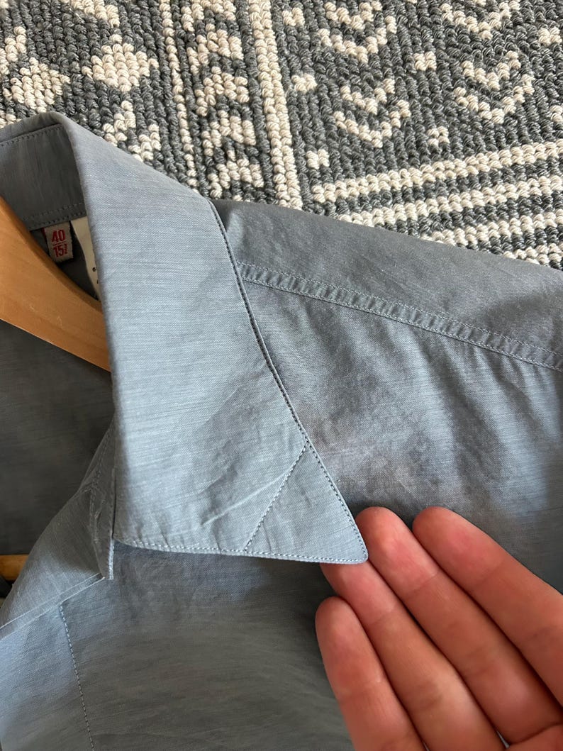 May include: Close-up of a light blue button-down shirt. The collar and part of the shirt are visible, with a tag showing size 40/15. The shirt is hanging on a wooden hanger.