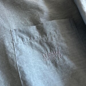 May include: Light blue button-up shirt with a chest pocket. The pocket features embroidered text that reads "VERSACE CLASSIC V2". The shirt is made of a lightweight, semi-sheer fabric.