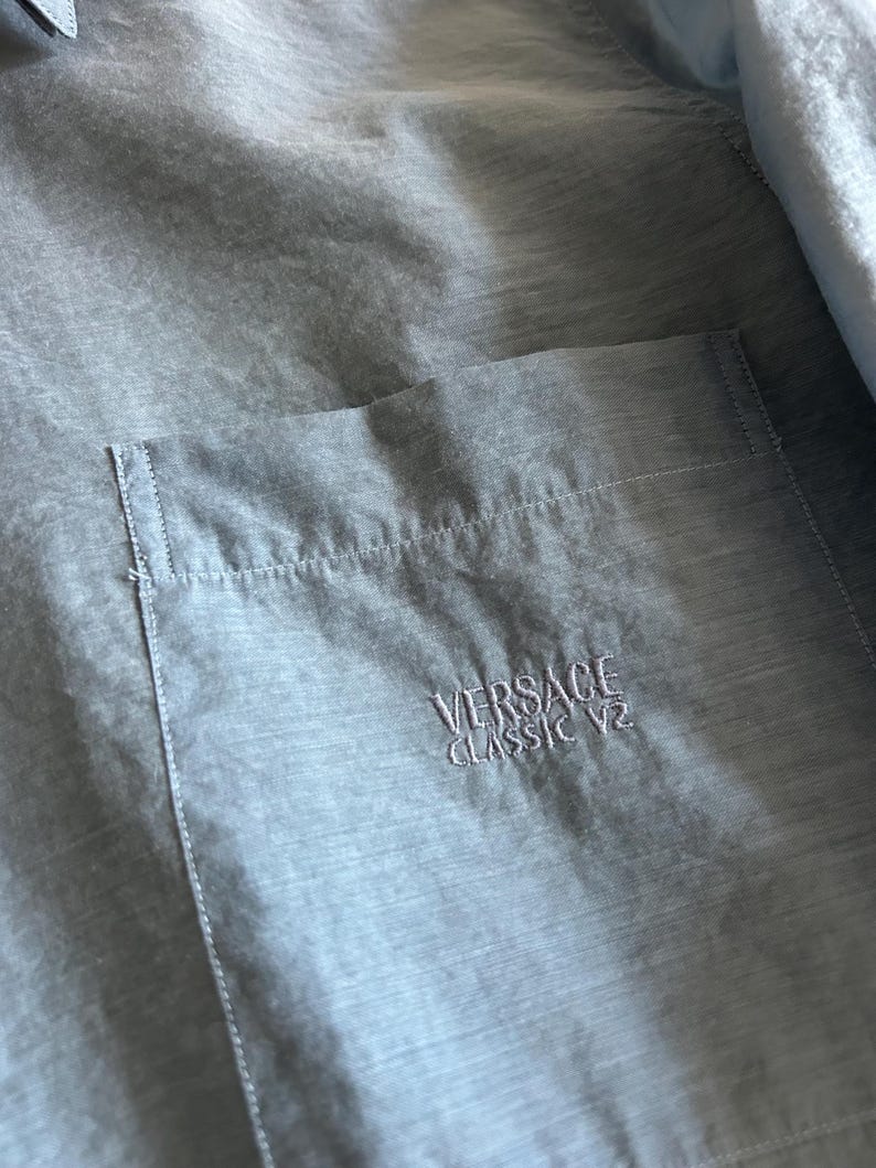 May include: Light blue button-up shirt with a chest pocket. The pocket features embroidered text that reads "VERSACE CLASSIC V2". The shirt is made of a lightweight, semi-sheer fabric.