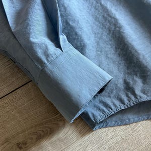 May include: Close-up of a light blue long-sleeved shirt. The cuff of the sleeve is visible, showing a tailored design with a button closure. The fabric has a subtle sheen, suggesting a smooth, possibly silky texture.