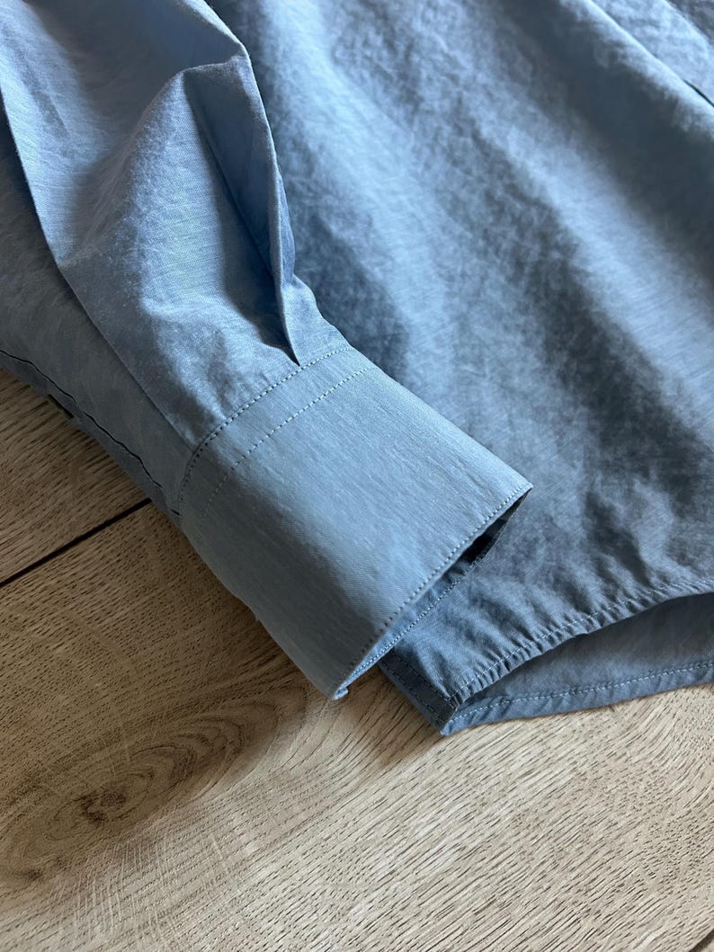 May include: Close-up of a light blue long-sleeved shirt. The cuff of the sleeve is visible, showing a tailored design with a button closure. The fabric has a subtle sheen, suggesting a smooth, possibly silky texture.