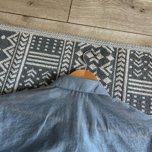 May include: A light blue collared shirt hanging on a wooden hanger. The shirt is made of a lightweight, semi-transparent fabric. The background includes a patterned rug and a wooden floor.
