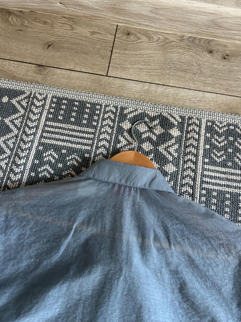 May include: A light blue collared shirt hanging on a wooden hanger. The shirt is made of a lightweight, semi-transparent fabric. The background includes a patterned rug and a wooden floor.