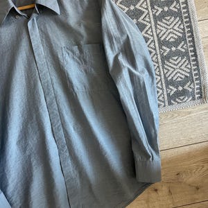 May include: A light blue button-down shirt with long sleeves, a collar, and a chest pocket. The shirt is made of a lightweight fabric and is hanging on a wooden hanger. The shirt has a subtle sheen and a classic design.
