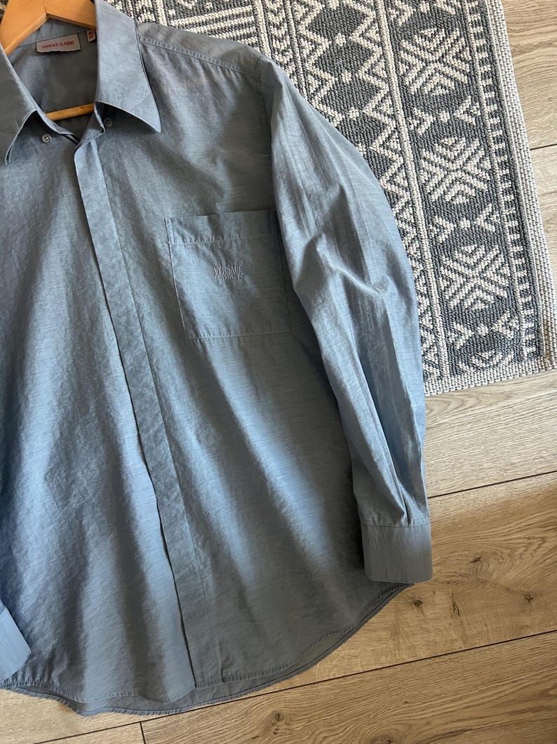 May include: A light blue button-down shirt with long sleeves, a collar, and a chest pocket. The shirt is made of a lightweight fabric and is hanging on a wooden hanger. The shirt has a subtle sheen and a classic design.