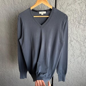 May include: A slate blue V-neck sweater with long sleeves, hanging on a wooden hanger. The sweater has ribbed cuffs and a ribbed hem. The brand name is visible on the neckline.