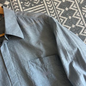 May include: A light blue button-down shirt with a chest pocket. The shirt has a classic collar and long sleeves. The Versace Classic V2 logo is embroidered on the pocket. The shirt is hanging on a wooden hanger.