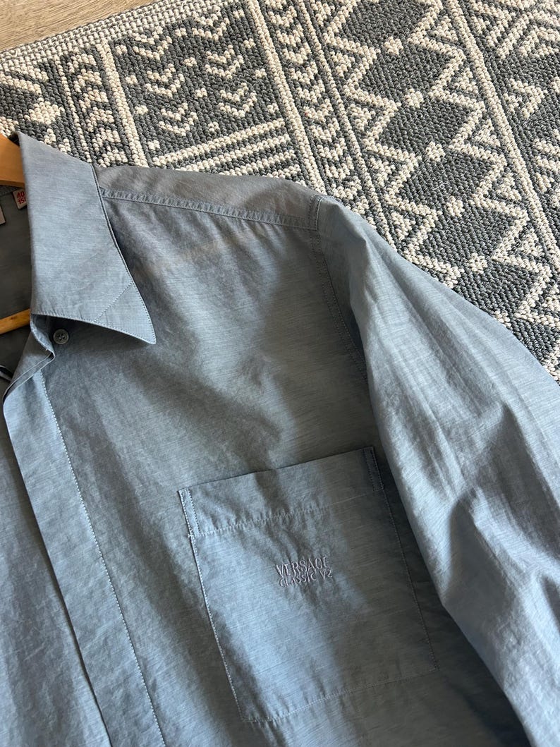 May include: A light blue button-down shirt with a chest pocket. The shirt has a classic collar and long sleeves. The Versace Classic V2 logo is embroidered on the pocket. The shirt is hanging on a wooden hanger.