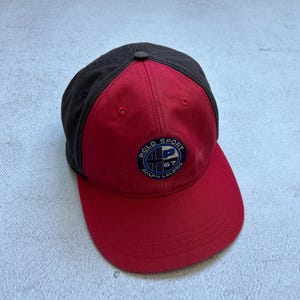May include: A red and black baseball cap with the text "POLO SPORT RALPH LAUREN" on the front. The cap has a curved brim and a button on top. The hat is made of a durable material.