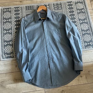 May include: A light blue long-sleeve shirt with a button-down front and a chest pocket. The shirt is made of a smooth fabric and is displayed on a wooden hanger. The shirt is laying on a patterned rug.