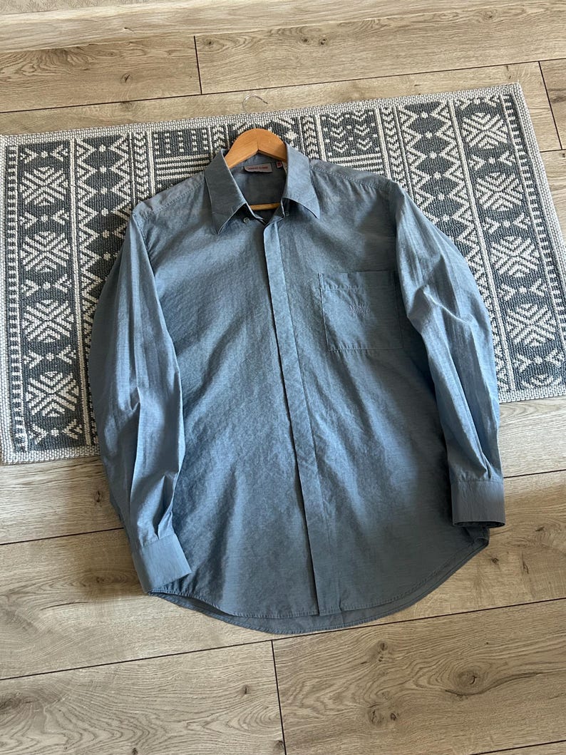 May include: A light blue long-sleeve shirt with a button-down front and a chest pocket. The shirt is made of a smooth fabric and is displayed on a wooden hanger. The shirt is laying on a patterned rug.