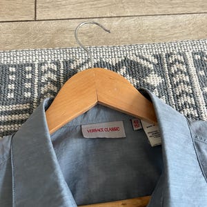 May include: A light blue button-down shirt with a collar, hanging on a wooden hanger. The label reads "VERSACE CLASSIC" in a red rectangle. The shirt is made of a smooth, possibly silk-like material.