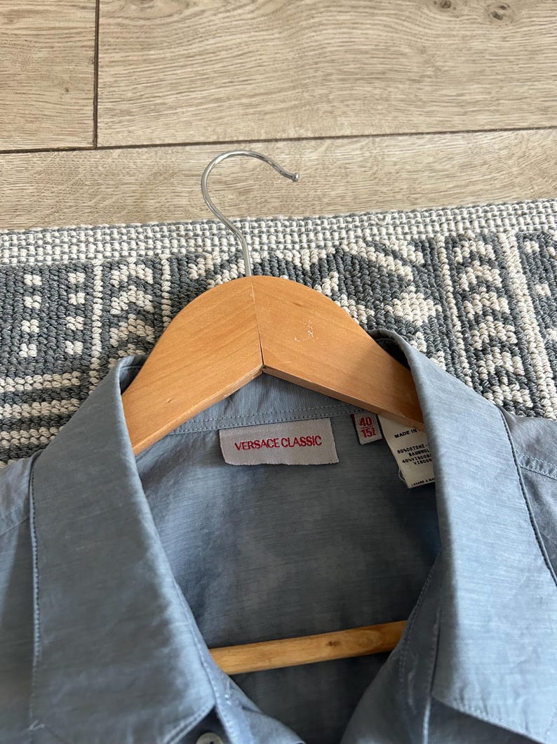 May include: A light blue button-down shirt with a collar, hanging on a wooden hanger. The label reads "VERSACE CLASSIC" in a red rectangle. The shirt is made of a smooth, possibly silk-like material.