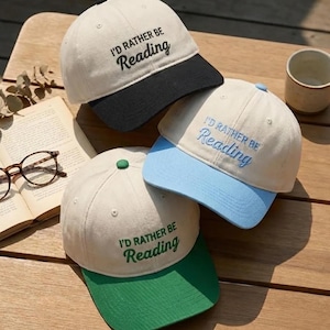 May include: Three baseball caps with the text "I'd Rather Be Reading". The caps have different color combinations: cream with a black brim, cream with a light blue brim, and cream with a green brim. The caps are on a wooden surface.